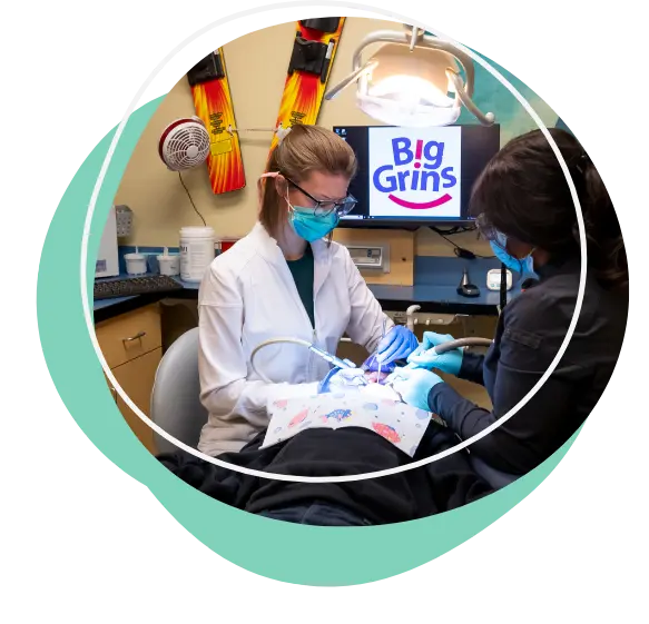 SpecializedEmergency Two dental professionals wearing masks and gloves work on a patient in a dental office. A screen in the background displays the "Big Grins" logo, and colorful skis are mounted on the wall.